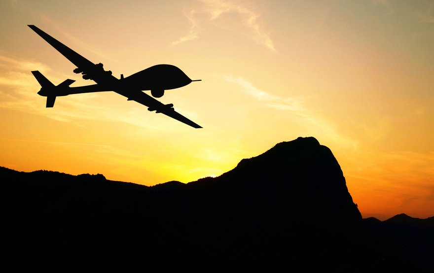 Military drone in the sky at sunset with sun rising over mountain in background.